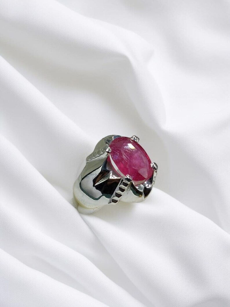 K&ouml;nnte beinhalten: Ein silberner Ring mit einem gro&szlig;en, ovalen, rosafarbenen Edelstein, der in einer Krappenfassung sitzt. Der Ring hat ein detailliertes Band mit einem strukturierten Design.