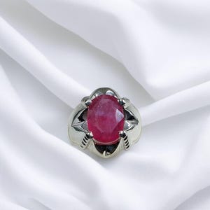 K&ouml;nnte beinhalten: Ein silberner Ring mit einem gro&szlig;en, ovalen Rubin-Edelstein. Der Ring hat eine Krappenfassung und eine strukturierte Band.