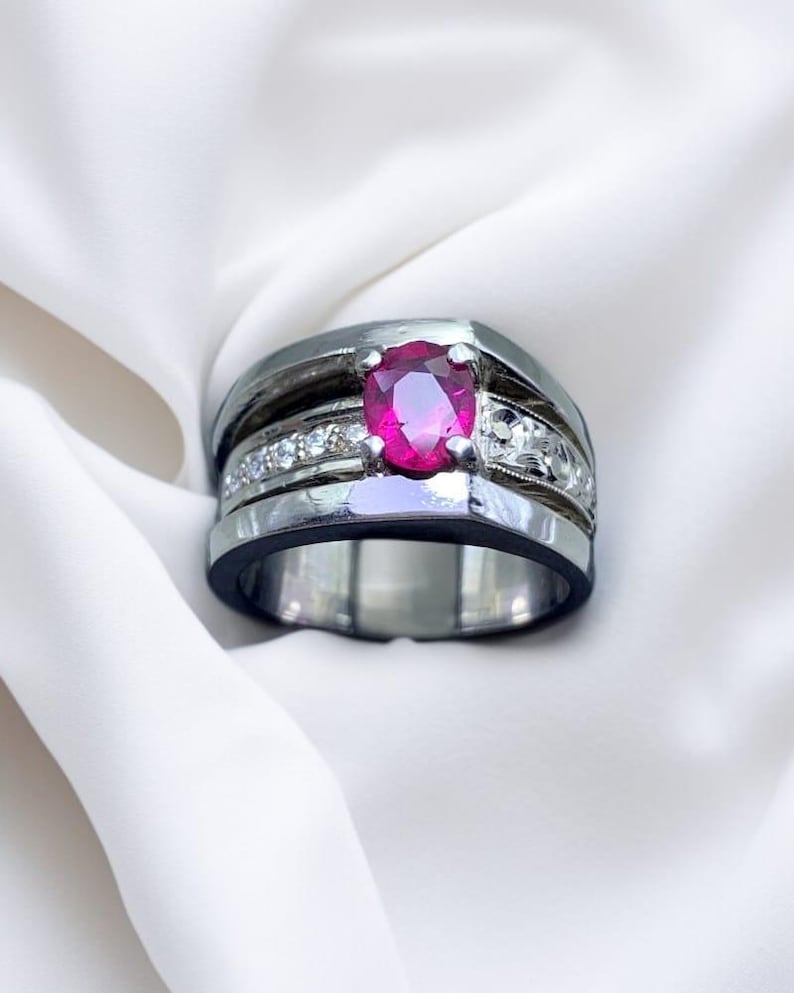 K&ouml;nnte beinhalten: Ein silberner Ring mit einem gro&szlig;en, ovalen Rubin-Edelstein, der von kleineren Diamanten umgeben ist. Der Ring hat ein breites Band mit einer schwarzen Einlage.