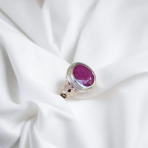 Op de afbeelding: Een zilveren ring met een grote, ovale robijn. De ring heeft ingewikkelde details rond de steen en de band.