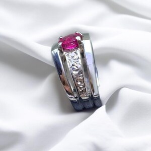 K&ouml;nnte beinhalten: Ein silberner Ring mit einem rosafarbenen Edelstein in der Mitte. Die Band ist mit einem floralen Muster graviert.