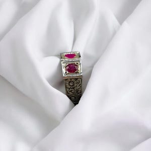 Op de afbeelding: Een zilveren ring met ingewikkelde bloemmotieven en twee rode edelstenen.