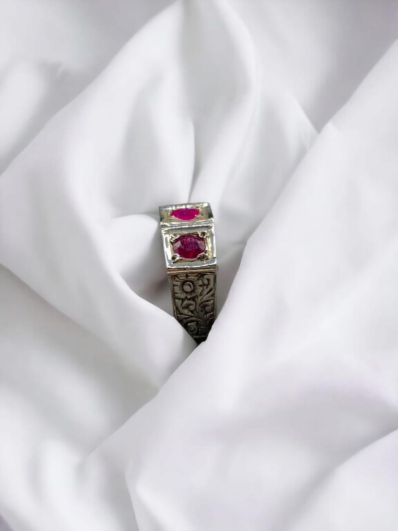 Op de afbeelding: Een zilveren ring met ingewikkelde bloemmotieven en twee rode edelstenen.