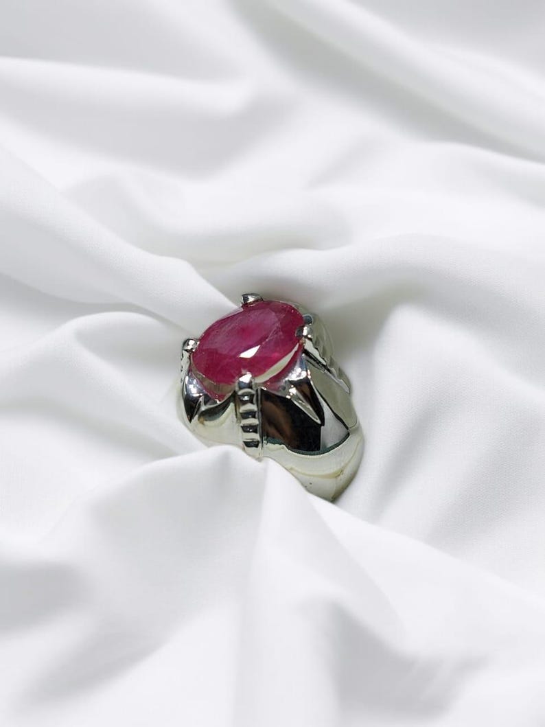 K&ouml;nnte beinhalten: Ein silberner Ring mit einem gro&szlig;en, ovalen Rubin-Edelstein. Der Ring hat eine detaillierte, strukturierte Band.