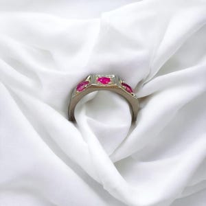 Op de afbeelding: Een gouden ring met vier rode edelstenen die in een vierkante vorm zijn gezet. De ring ligt op een witte stoffen achtergrond.