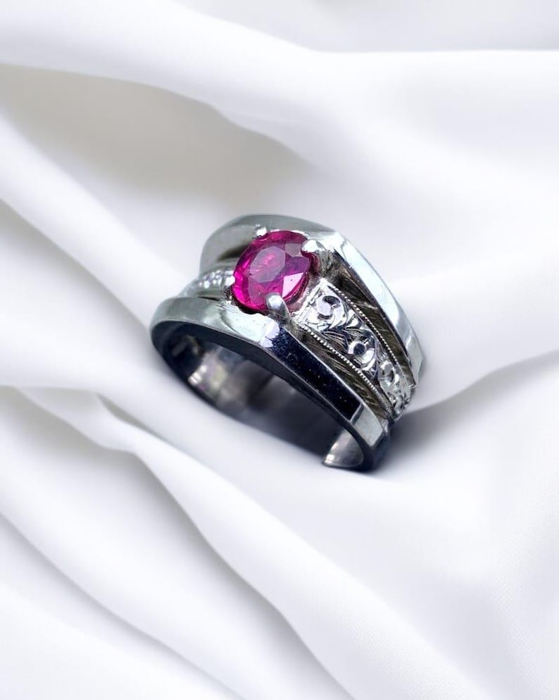 K&ouml;nnte beinhalten: Ein silberner Ring mit einem gro&szlig;en, ovalen Rubin-Edelstein, der in der Mitte eingesetzt ist. Der Ring hat eine dekorative, gravierte Band.