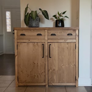 May include: A wooden cabinet with two doors and two drawers. The cabinet is stained a light brown colour and has black metal handles. The cabinet is sitting on a tiled floor in front of a white wall.