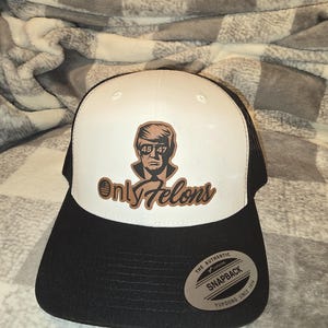 Op de afbeelding: Een witte en zwarte trucker-pet met een bruin leren patch waarop "Only Felons" staat en een afbeelding van een man met een zonnebril en het nummer "45 47" erop. De pet heeft een zwart mesh-achterpaneel en een zwarte drukknopsluiting. De pet heeft ook een bruin leren patch waarop "The Authentic Yupoong Snapback Since 1974" staat.