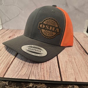 Könnte beinhalten: Graue Snapback-Kappe mit orangefarbenem Mesh-Rückenteil. Die Kappe hat einen braunen Lederaufnäher mit den Worten "Certified OSHA Violator" und dem Snapback-Logo.