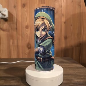 May include: A white cylindrical lamp base with a clear cylinder lampshade featuring a cartoon image of Link from the Legend of Zelda video game series. The image shows Link in a green tunic and cap, holding a sword, with a blue background.