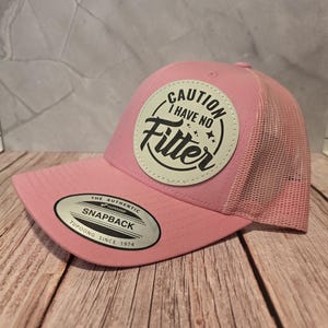 Puede incluir: Gorra rosa con parte trasera de malla y visera curva. La gorra presenta un parche circular con el texto "CAUTION I HAVE NO Filter" en negro. Cierre trasero ajustable y etiqueta "Yupoong".