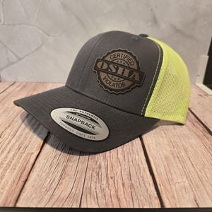 Könnte beinhalten: Graue und neongelbe Snapback-Kappe mit gebogenem Schirm. Die Kappe hat einen Lederaufnäher mit den Worten "CERTIFIED OSHA VIOLATOR" und der Marke "Yupoong" seit 1974.