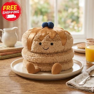 May include: A plush pancake toy with a smiling face, syrup, and two blueberry toppings. The pancake is sitting on a white plate with two small feet. The toy is made of soft, textured material.