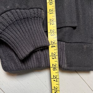 May include: Close-up of a dark grey jacket sleeve with a ribbed cuff. A yellow measuring tape shows the jacket's length is approximately 61 cm. The jacket appears to be made of a thick, possibly wool, material.