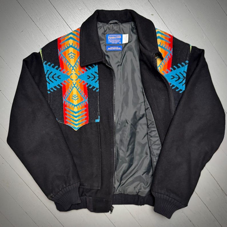 May include: Black jacket with a vibrant geometric pattern in blue, orange, and yellow on the shoulders and chest. It has a zip closure and a collar. The interior lining is grey. The jacket's design is reminiscent of traditional textiles.