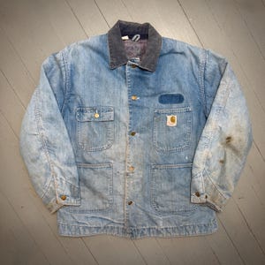 May include: Light blue denim jacket with a dark grey corduroy collar. The jacket features four front pockets, brass buttons, and a Carhartt logo patch. The jacket shows signs of wear, with some staining on the sleeves.