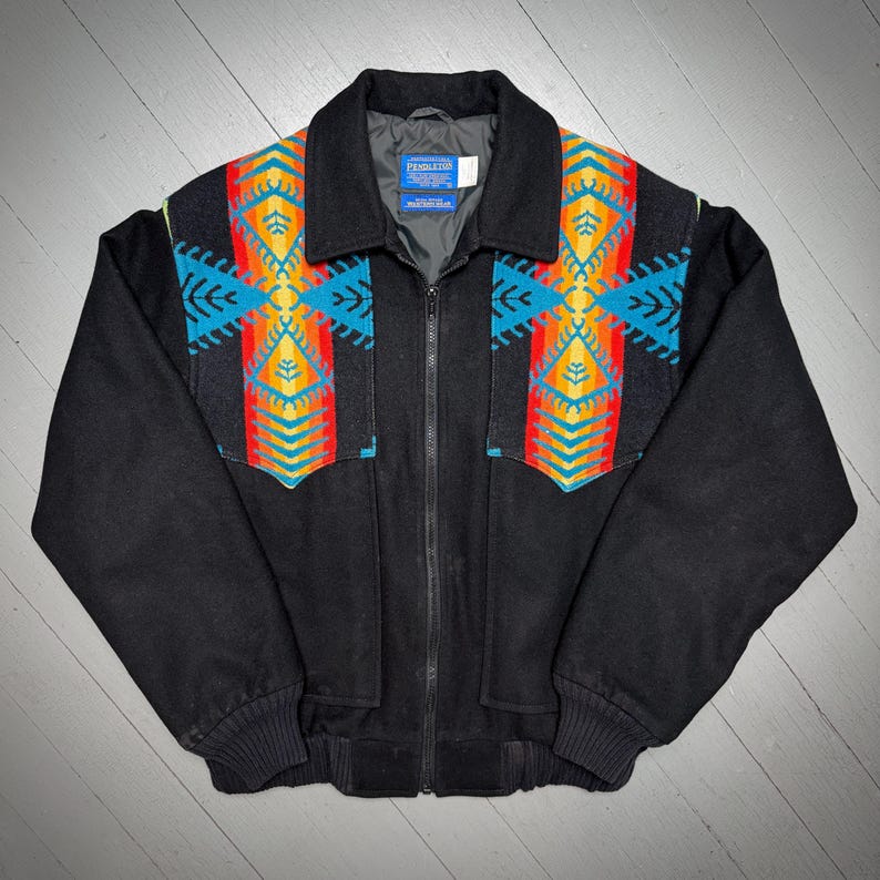 May include: Black zip-up jacket with colourful geometric patterns on the chest and shoulders. The jacket has a collar, two front pockets, and ribbed cuffs. The Pendleton label is visible on the inside.