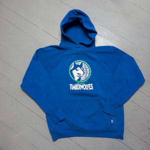 May include: A blue hooded sweatshirt with the Minnesota Timberwolves logo. The logo features a wolf's head, a basketball, and the text "Timberwolves". The sweatshirt has a front pocket and a hood.