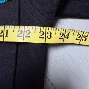 May include: A dark navy blue garment is measured with a yellow measuring tape. The tape shows measurements from 53.3 to 63.5 cm. The garment appears to be made of a thick fabric.