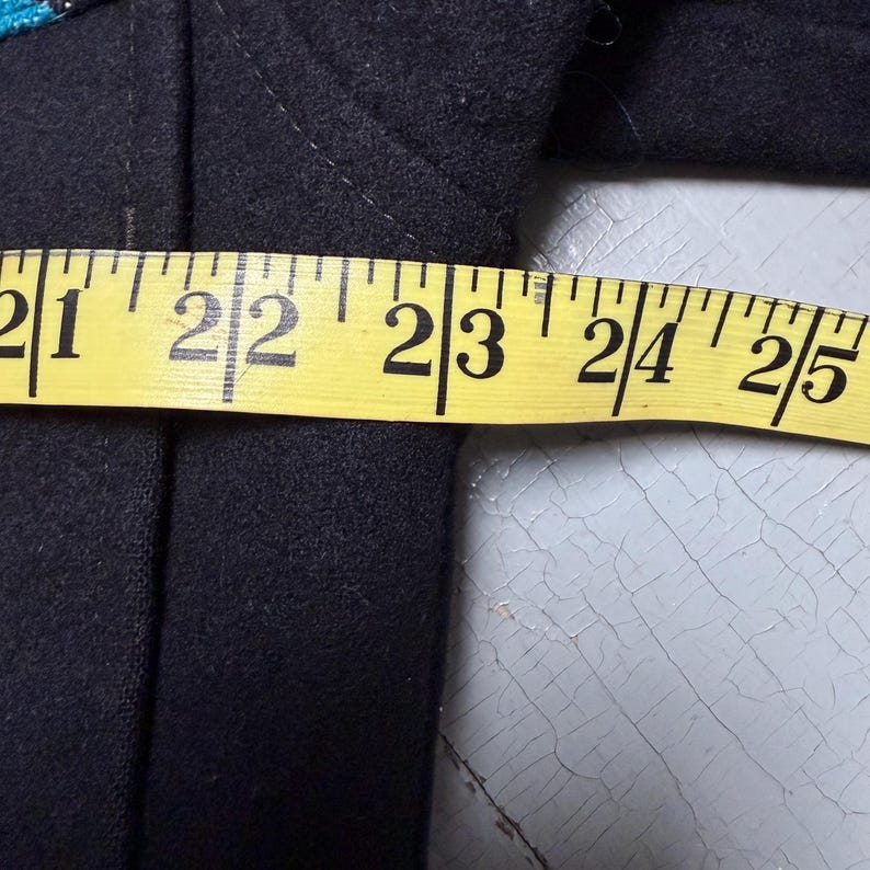 May include: A dark navy blue garment is measured with a yellow measuring tape. The tape shows measurements from 53.3 to 63.5 cm. The garment appears to be made of a thick fabric.