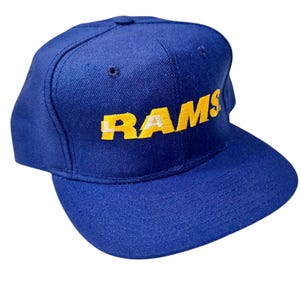 May include: A blue baseball cap with the word "RAMS" in yellow block letters across the front. The hat has a curved brim and a structured crown. The hat is a solid blue color.