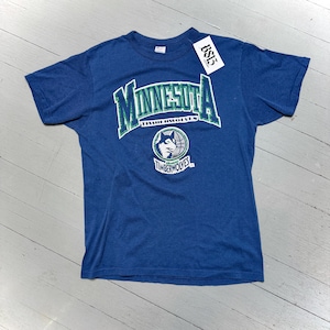 May include: A blue t-shirt featuring the word "MINNESOTA" in green and white lettering. Below the word "MINNESOTA" is the word "TIMBERWOLVES" in green lettering. A logo of a wolf inside a basketball is also on the shirt.