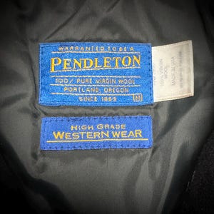 May include: A close-up of a black garment featuring three labels. The main blue label displays "PENDLETON" and "100% PURE VIRGIN WOOL". A second blue label reads "HIGH GRADE WESTERN WEAR". A white label states "100% VIRGIN WOOL MADE IN USA".