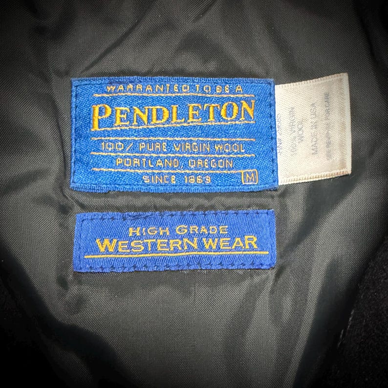 May include: A close-up of a black garment featuring three labels. The main blue label displays "PENDLETON" and "100% PURE VIRGIN WOOL". A second blue label reads "HIGH GRADE WESTERN WEAR". A white label states "100% VIRGIN WOOL MADE IN USA".