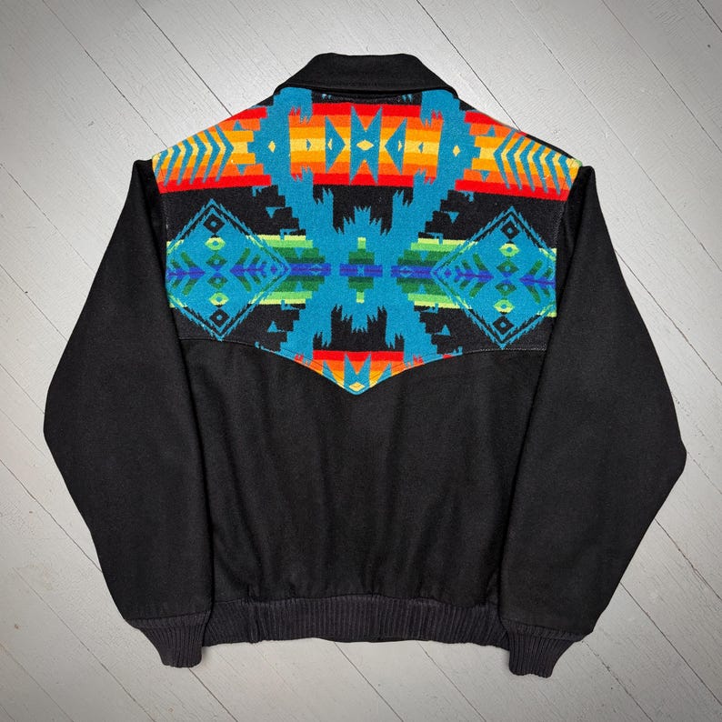 May include: Black jacket featuring a vibrant geometric design in blue, red, orange, and yellow on the back. The jacket has a collar and ribbed cuffs and hem. A stylish outerwear piece.
