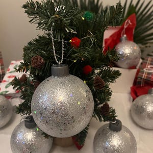 May include: A small artificial Christmas tree decorated with silver glitter ornaments. The tree has pinecones and red and green decorative accents. Several additional silver ornaments are arranged around the base of the tree, creating a festive holiday display.