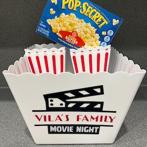 May include: A white popcorn bucket with red and white stripes around the top. The bucket has a black and white movie clapper design and the text "VILA'S FAMILY MOVIE NIGHT". A box of Pop Secret popcorn with the text "EXTRA BUTTER" is on top of the bucket.