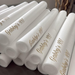 Può includere: Una collezione di tubi cilindrici in schiuma bianca, ciascuno con il testo Gabby's XV stampato in un carattere corsivo color oro metallizzato. Questi bastoncini di schiuma cavi sono disposti in una formazione impilata, evidenziando le scritte.