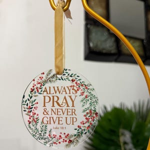 以下が含まれることがあります： 「ALWAYS PRAY & NEVER GIVE UP」の金色の文字が書かれた、緑の葉と赤い実のリースで囲まれた透明な丸いオーナメント。ベージュのリボンが付いた金色のフックから吊り下げられています。