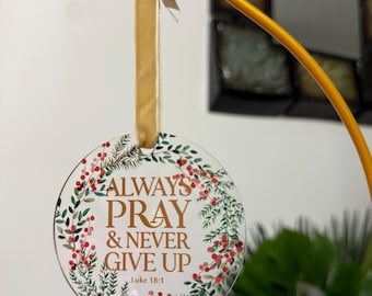 May include: A clear, circular ornament featuring the gold text "ALWAYS PRAY & NEVER GIVE UP", encircled by a wreath of green foliage and red berries. It is suspended from a gold hook with a beige ribbon.
