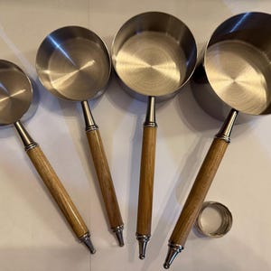 May include: A set of four stainless steel measuring cups with long, cylindrical wooden handles. The cups vary in size from smallest to largest, each featuring a polished metal interior and a sturdy, light-toned wood grip with metal accents.