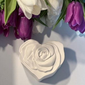 May include: A white ceramic heart-shaped rose sculpture. The rose is detailed with multiple layers of petals.