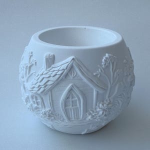 May include: A white, spherical ceramic planter with a detailed relief design of a fairy house. The house features a pitched roof, arched door, and windows, surrounded by floral and foliage elements. The planter has a wide opening.