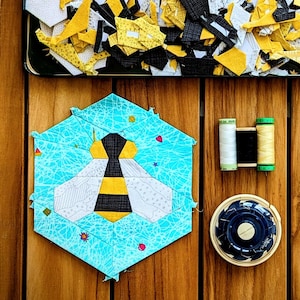 May include: A blue and white patchwork bee quilt block with a yellow and black bee design. The block is surrounded by scraps of fabric in various colors and patterns, including yellow, white, and black. The scraps are arranged in a tray with a wooden background.