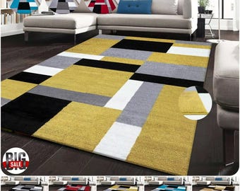 May include: A large, rectangular area rug with a geometric pattern. The rug features blocks of yellow, black, gray, and white. The rug is displayed in a living room setting, with a sofa and side table visible in the background. Several color variations are shown.