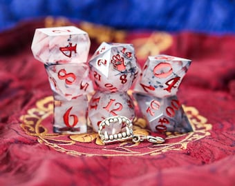 May include: A set of six clear, multi-sided dice with red numbers and black and white swirls. The dice are arranged in a pyramid shape on a red and gold fabric. A silver charm with a set of fangs is in front of the dice.