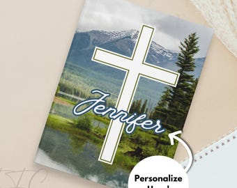 Op de afbeelding: Een notitieboek met een landschap, een wit kruis en de naam "Jennifer" in cursief. De afbeelding toont bergen, een meer en bomen. Een witte cirkel met de tekst "Personaliseer hier!" is ook aanwezig.