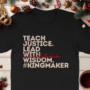 Peut inclure: T-shirt noir avec le texte blanc "TEACH JUSTICE. LEAD WITH Queen Bathsheba WISDOM. #KINGMAKER". Le t-shirt est entouré de décorations de Noël.