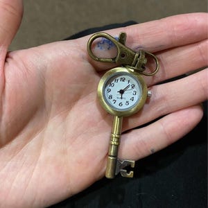 May include: A vintage-style key-shaped pocket watch with a quartz movement. The watch has a white face with black numerals and hands, and a brass-colored metal casing and key design. It includes a clasp for attaching to a bag or keychain.