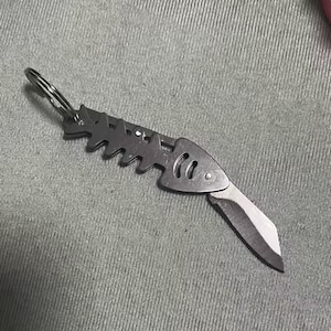 May include: A small, silver folding knife with a fish skeleton design. The blade is open, showing a sharp edge. The knife is attached to a key ring.