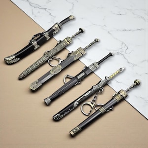 Può includere: Sei spade decorative in miniatura con manici e foderi ornati. Le spade sono in varie tonalità di argento, oro e nero. Alcune hanno portachiavi attaccati. Le spade sono disposte diagonalmente su una superficie di marmo e beige.