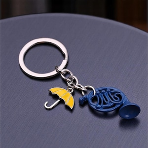 May include: A silver keychain with a yellow umbrella charm and a blue French horn charm. The umbrella has a curved handle and the horn has detailed lines. The keychain is on a dark surface.