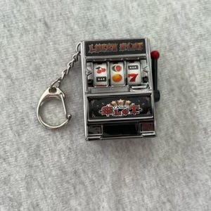 Może przedstawiać: Miniaturowy brelok do kluczy w kształcie automatu do gier z napisem "LUCKY SLOT" u g&oacute;ry i "CASINO SLOT" na dole. Na bębnach widać wiśnie, pomarańczę i cyfrę 7. Uchwyt jest czerwony. Brelok jest srebrny.