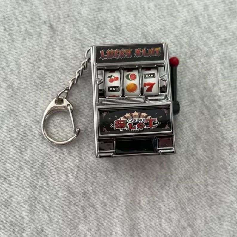 Może przedstawiać: Miniaturowy brelok do kluczy w kształcie automatu do gier z napisem "LUCKY SLOT" u g&oacute;ry i "CASINO SLOT" na dole. Na bębnach widać wiśnie, pomarańczę i cyfrę 7. Uchwyt jest czerwony. Brelok jest srebrny.