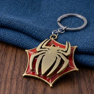 May include: A gold-colored metal keychain featuring a spider emblem. The spider is set against a red web design, with a silver key ring attached. The keychain is a superhero-themed accessory.