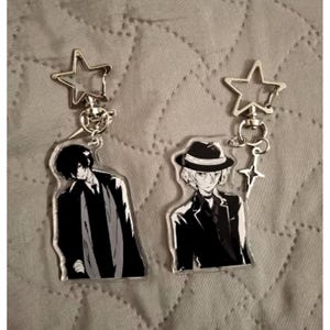 Peut inclure: Deux porte-clés de personnages de style anime. L'un présente un personnage en manteau et cravate noirs, l'autre un personnage en fedora et costume. Les deux porte-clés ont un dessus en forme d'étoile et un petit charme en forme d'étoile.